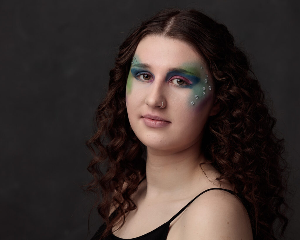 A person with long, wavy dark brown hair faces the camera against a dark grey background. They wear artistic makeup featuring blue and green eyeshadow with pink accents, and small silver studs along their cheekbone. They wear a black sleeveless top and have a neutral expression.