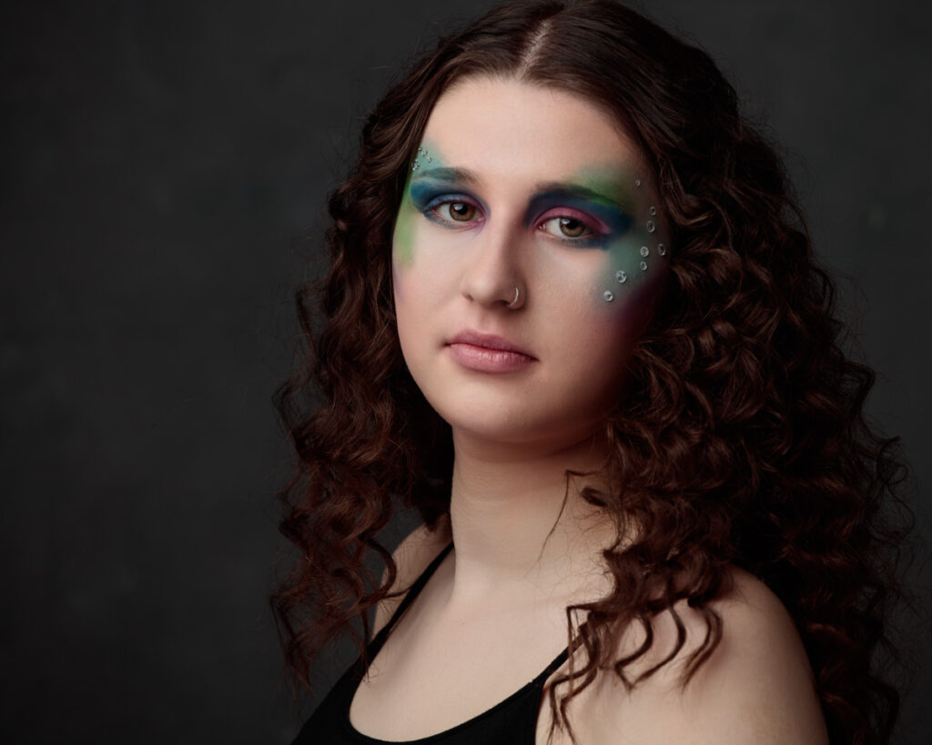 A person with long, wavy dark brown hair and artistic eye makeup in shades of green, blue, and pink against a dark background.