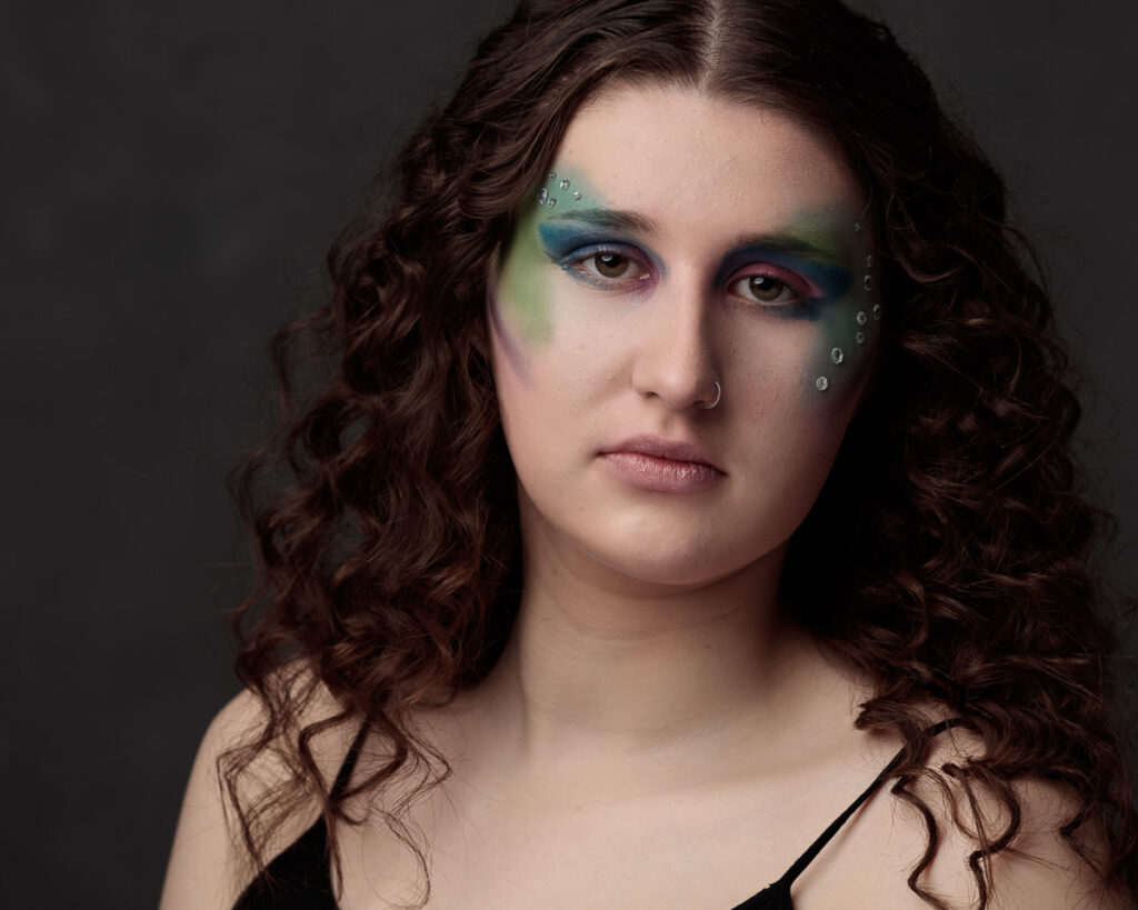 Portrait of a person with long curly brown hair wearing artistic eye makeup in blue, green and pink tones against a dark background.