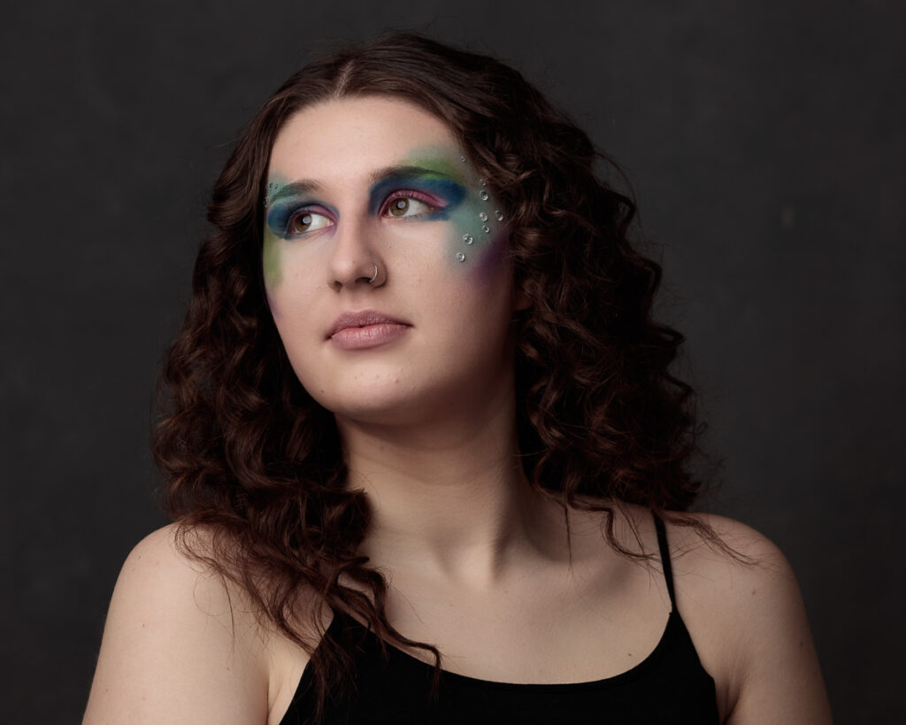 Portrait of a person with long dark curly hair wearing artistic eye makeup in blue, green, and purple tones with decorative rhinestones on their cheek, against a dark grey background.