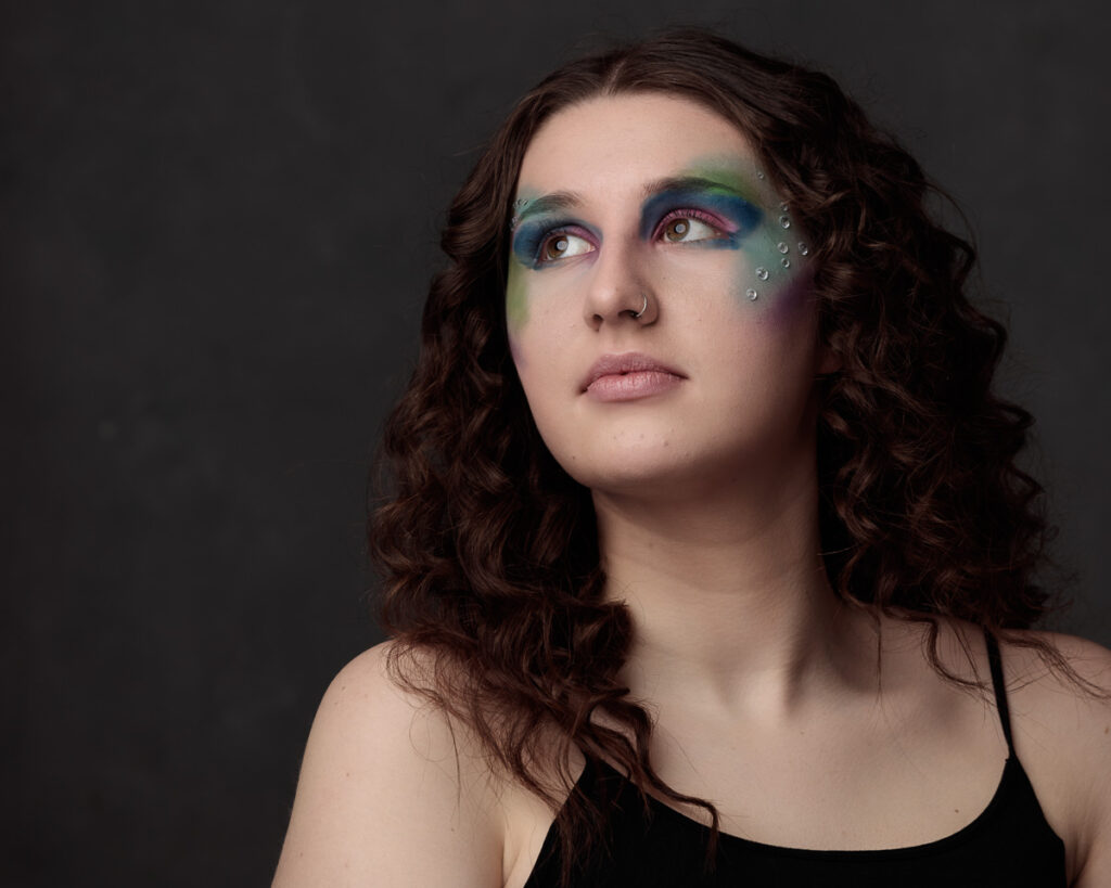 A person with shoulder-length dark wavy hair wears vibrant eyeshadow in blue, green, and purple tones with small silver rhinestones below one eye against a dark grey background.
