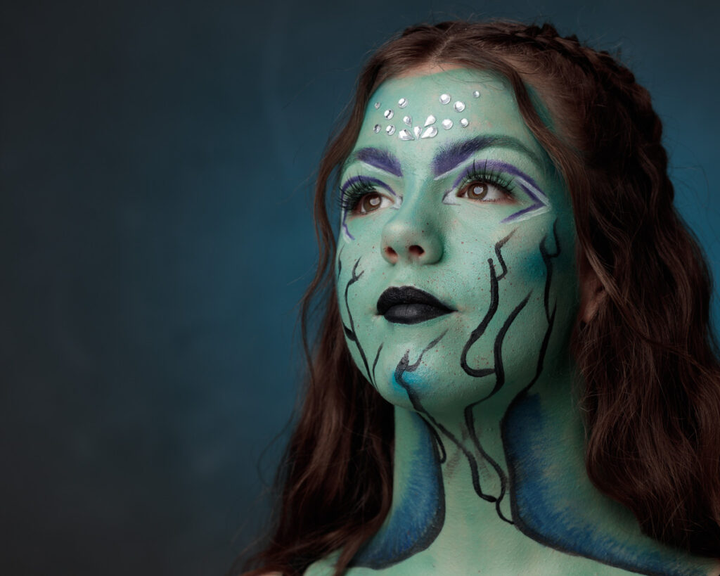 A woman with green face paint, purple eyeshadow, black lips, and decorative gems on her forehead against a blue background.