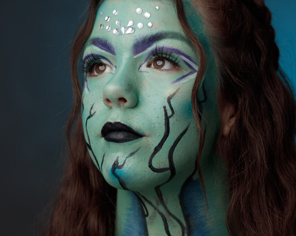 A woman with long brown hair wears elaborate face paint in shades of green and blue, with purple eyeshadow, black lips, and decorative silver studs on her forehead.