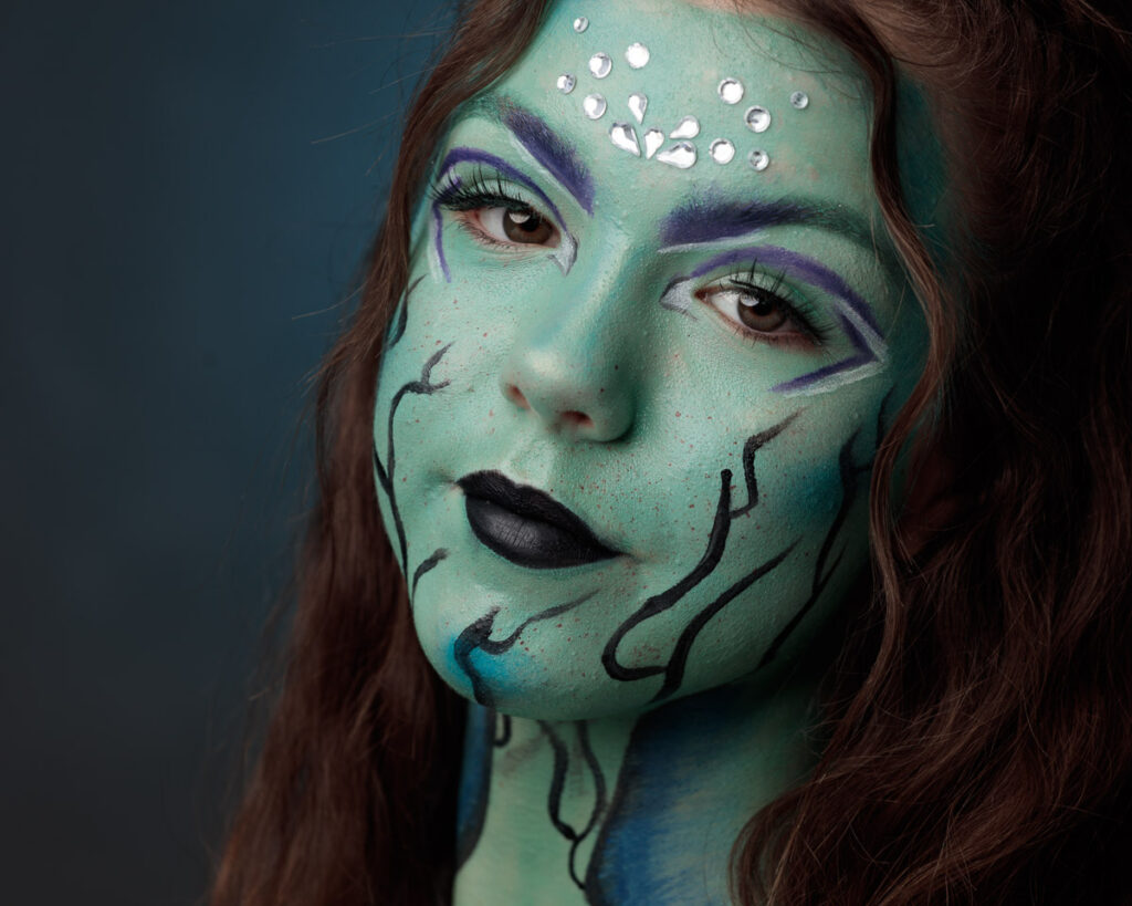 A person with green face paint, purple eyeshadow, black lips, and decorative silver studs on their forehead against a dark blue background.