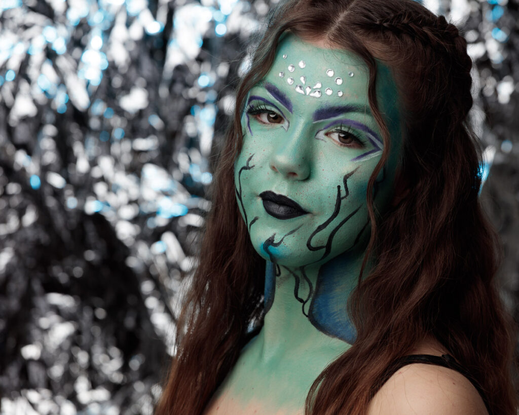 A woman with long brown hair wearing elaborate green face paint with purple eyebrows, black lips, and decorative gems, against a blurred sparkly background.
