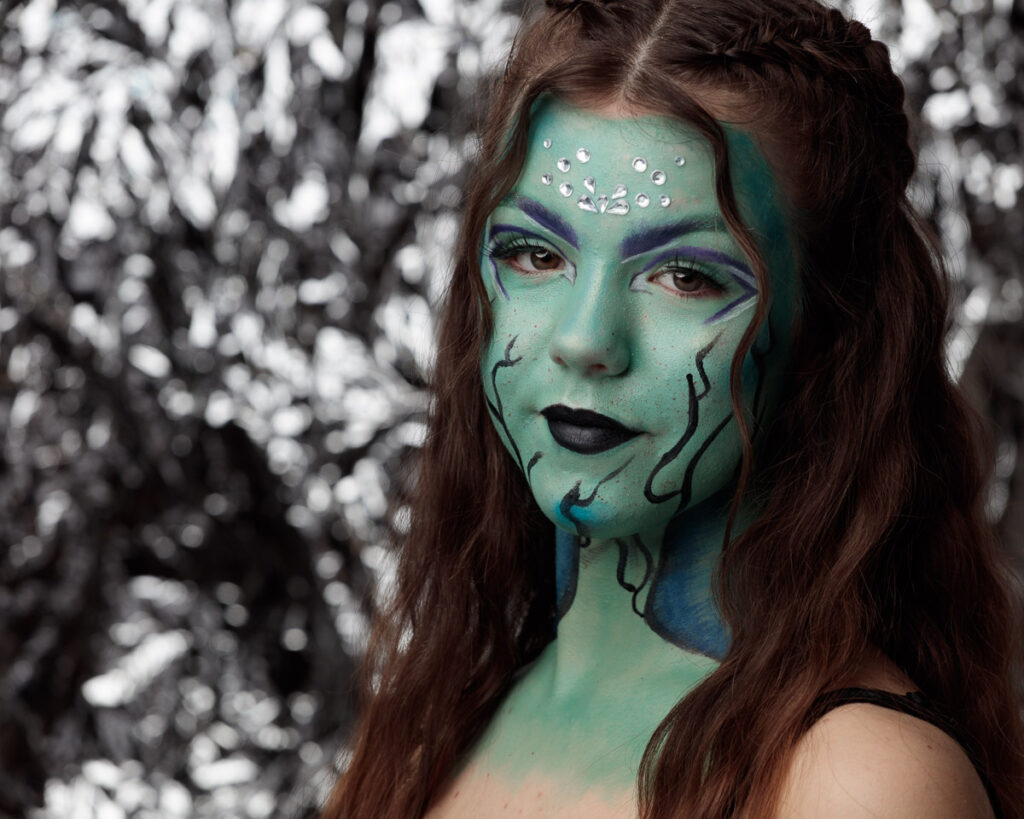 Woman with green face paint and decorative rhinestones, dark eyeshadow, black lips, against blurred snowy foliage background.