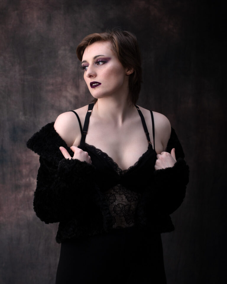 A woman in a black lace slip dress and fuzzy shrug looks to the side against a dark textured background.