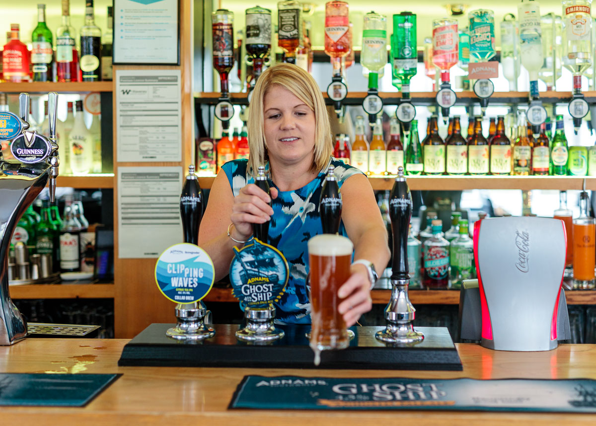 A woman pours a pint of beer at a bar with multiple beer taps and bottles displayed on shelves behind her.