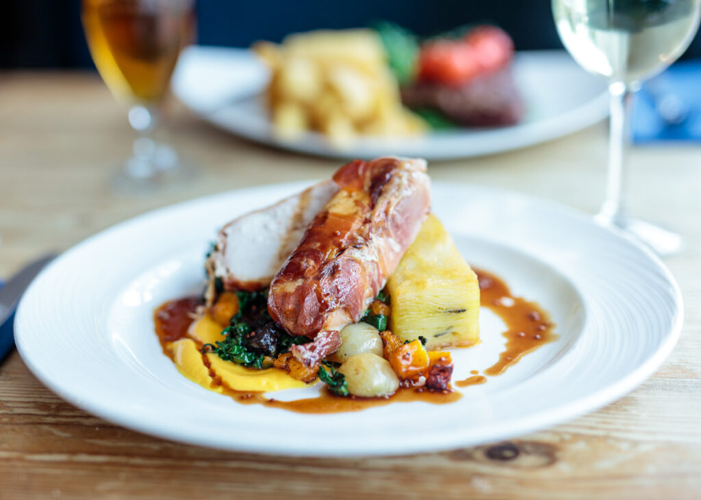 A seared duck breast served on polenta with dark leafy greens and root vegetables, drizzled with sauce, on a white plate.