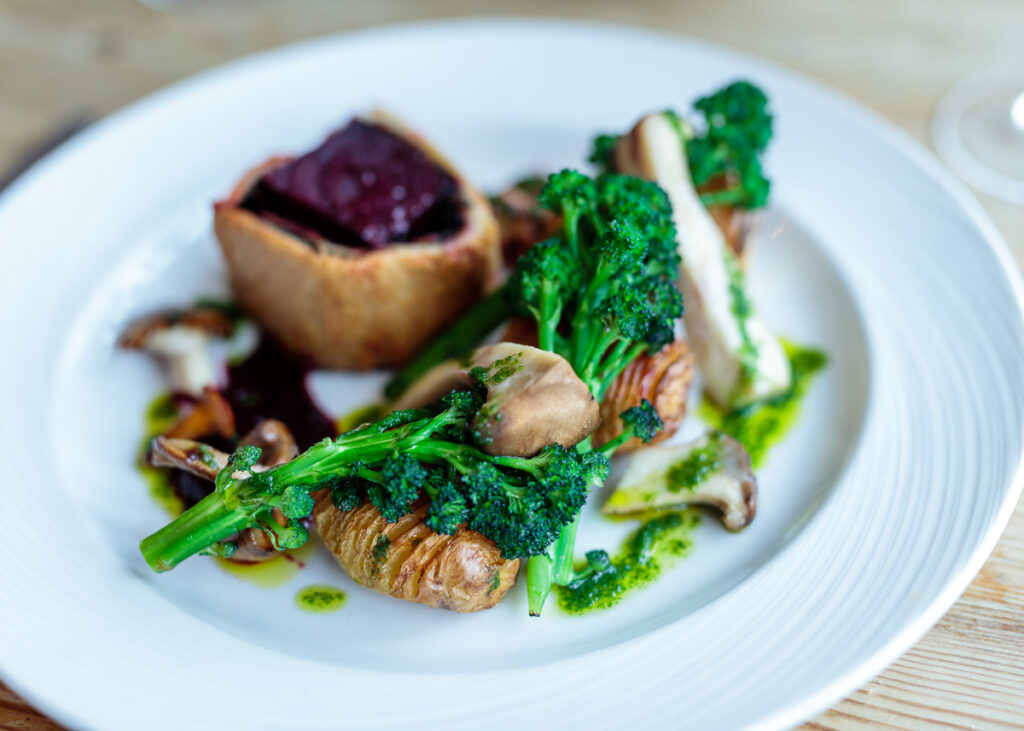 A plated dish featuring seared broccoli with mushrooms, charred meat, and a beetroot-filled pastry case on a white plate.