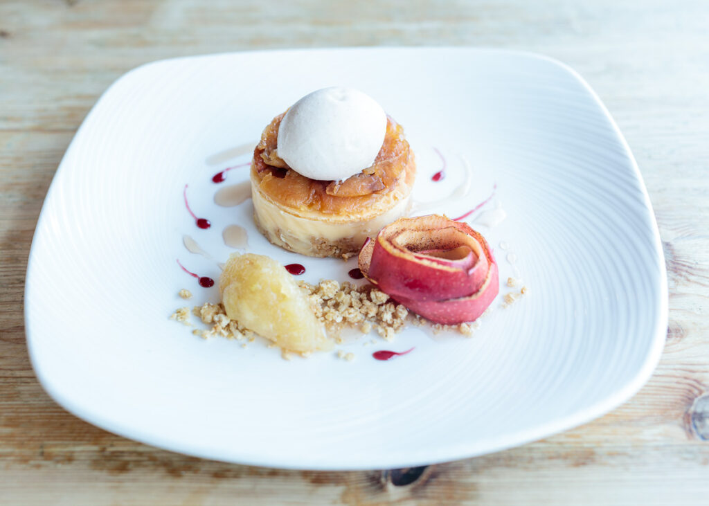 An elegant plated dessert on a white square plate featuring a pastry tower topped with meringue, a pink apple roll, crumble, and fruit coulis drizzle.