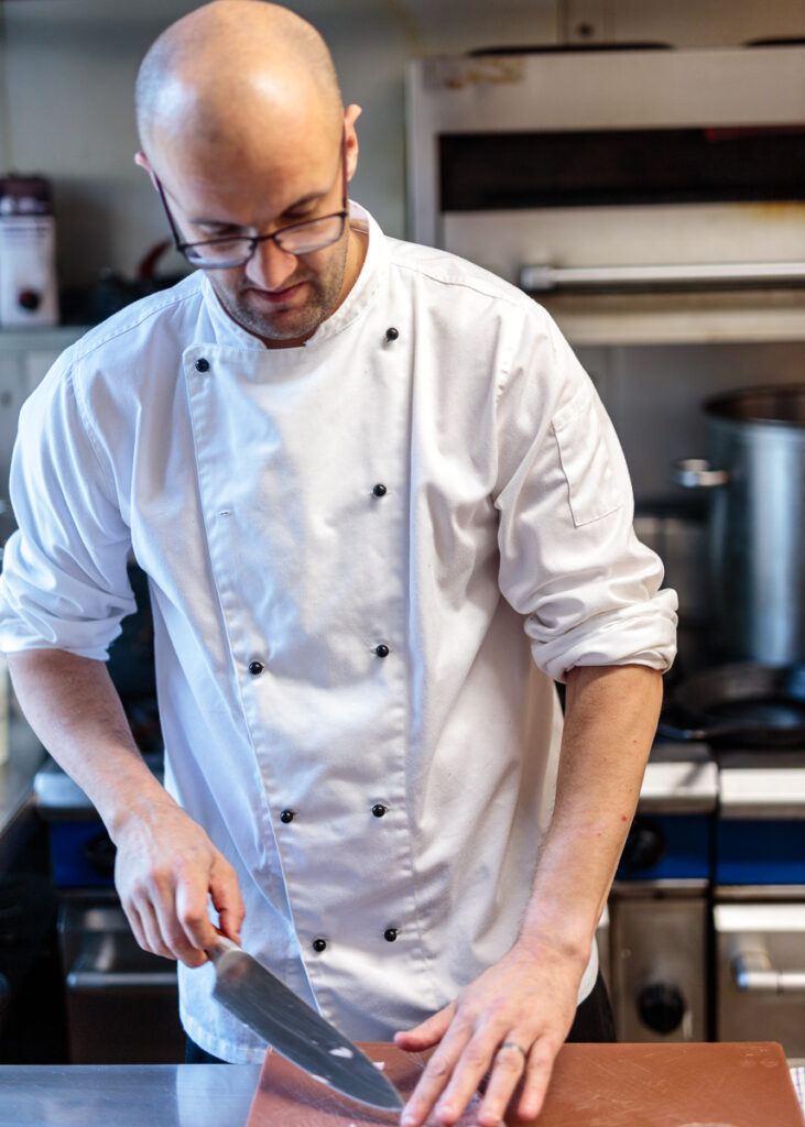 A chef in a white double-breasted jacket and glasses works at a counter in a professional kitchen.