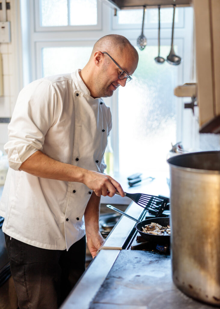 A man in a cream chef's shirt cooks at a stovetop in a bright kitchen, stirring food in a pan with a spatula.