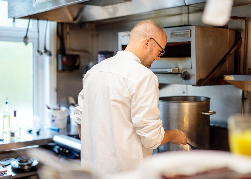 A man in a white shirt and glasses stands in a kitchen, working at a counter near a large metal pot.
