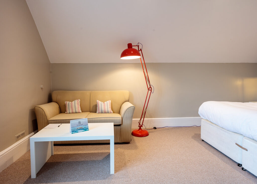 A modern hotel room with a yellow sofa, white table, red floor lamp, and bed against sloped ceiling.