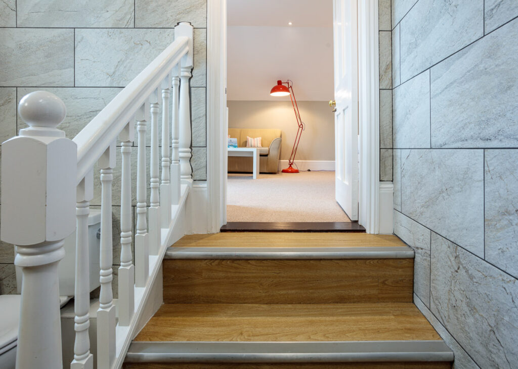 Staircase with white bannister descending to a hallway. Slate tiles line the walls. A red desk lamp is visible in the room ahead.