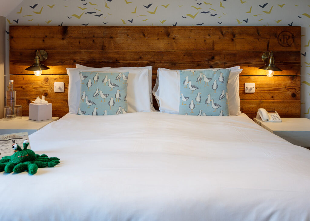 A hotel bedroom with a wooden headboard, white bedding, blue patterned cushions, bedside lamps, and a green toy octopus on the bed.