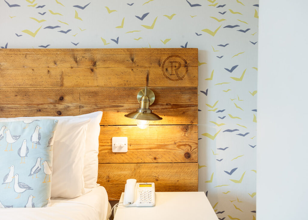 A hotel bedside with wooden headboard, brass sconce light, white pillow, phone, and patterned wallpaper with birds and chevrons.