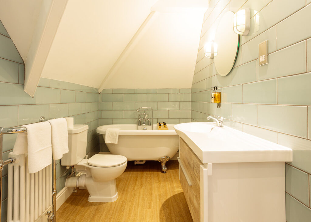 A spacious bathroom with pale green subway tiled walls, cream ceiling, wooden floor, freestanding bathtub, toilet, and double sink vanity.