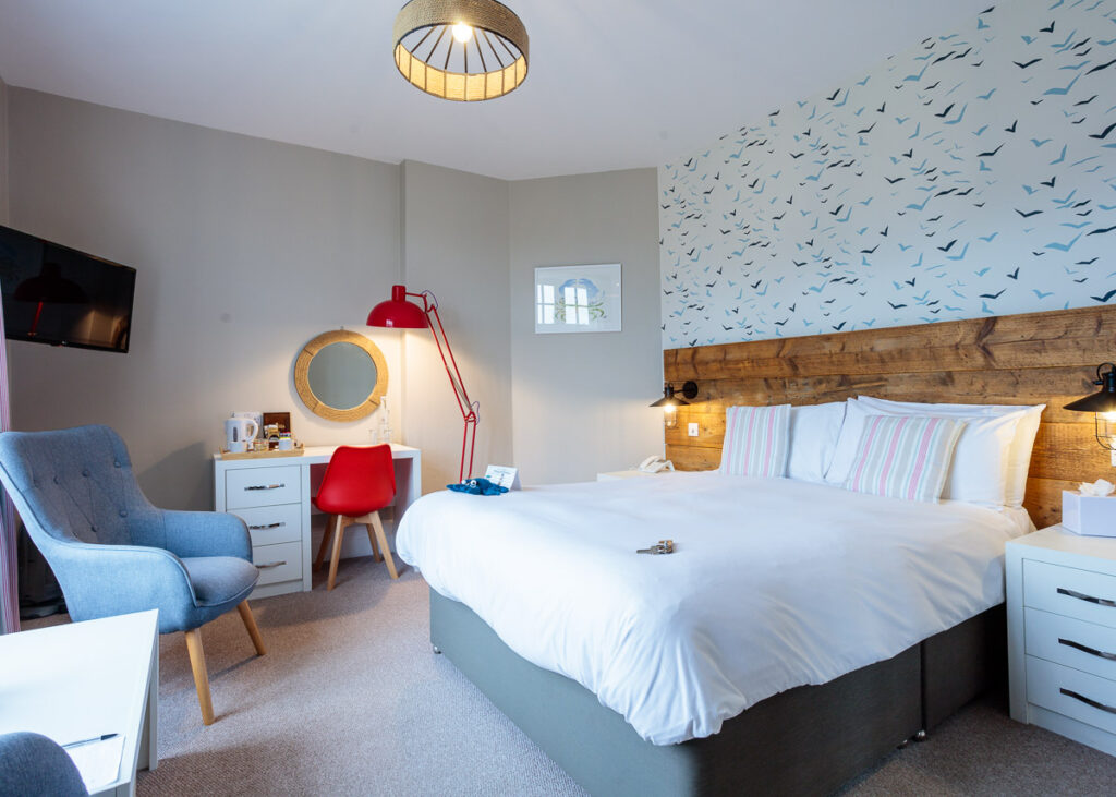 A modern hotel bedroom with a wooden headboard bed, blue accent chair, red desk lamp, and wallpaper featuring flying birds.