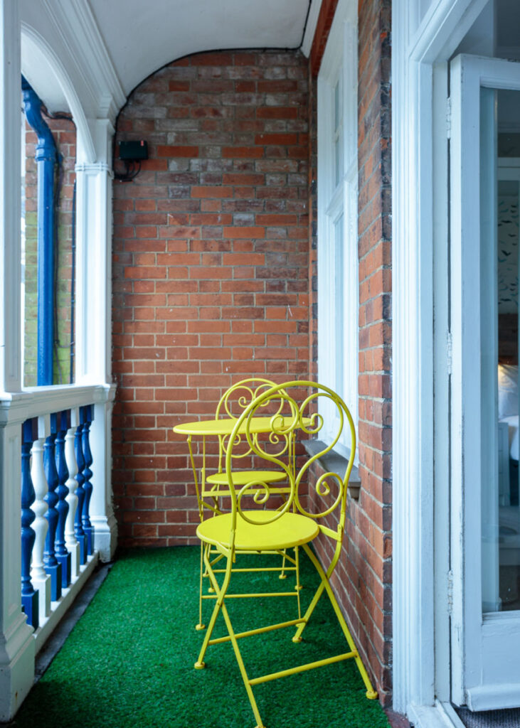 A bright yellow ornate metal chair sits on green artificial turf in a narrow brick passageway between white-trimmed walls.