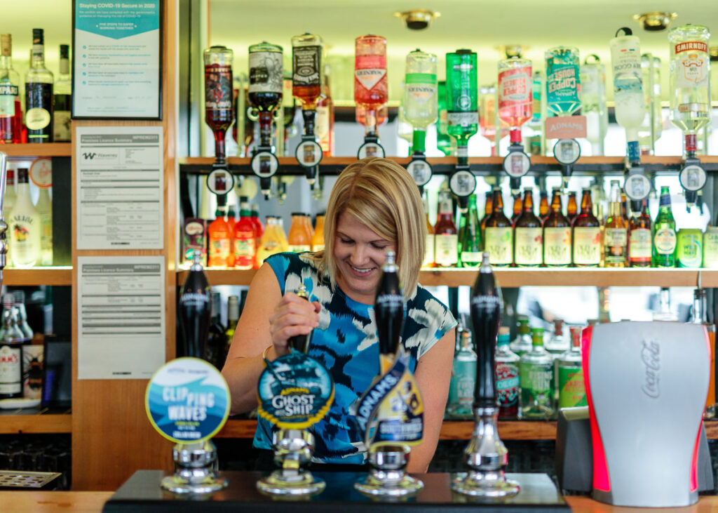 A woman stands behind a bar counter, smiling while adjusting a beer tap. Multiple spirits bottles line the shelves behind her.