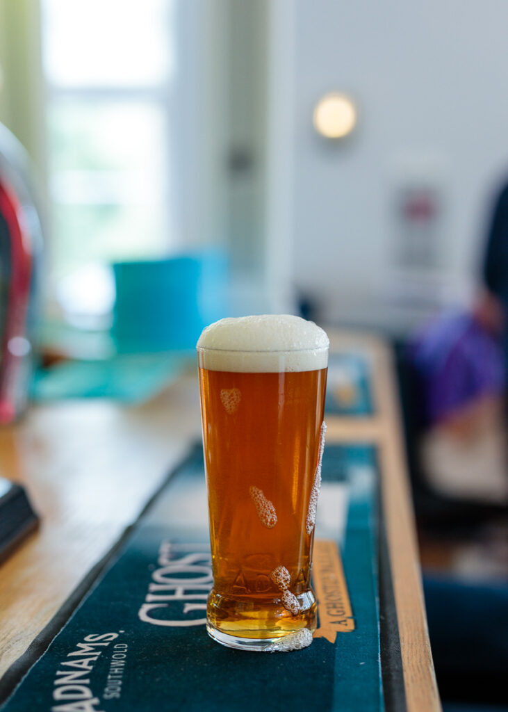 A pint glass of amber beer with a thick cream head sits on a bar mat in a pub, with a blurred interior background.
