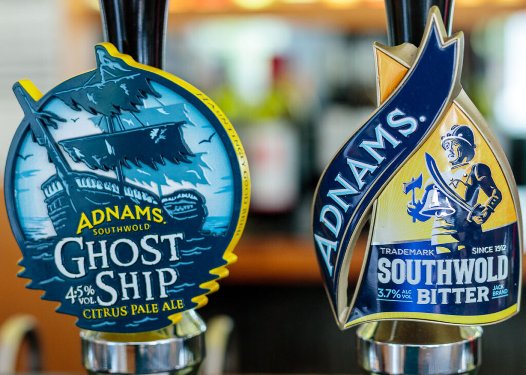 Two Adnams Southwold beer pump clips on a bar. Left shows Ghost Ship pale ale at 4.5% ABV with a ship illustration. Right shows Southwold Bitter at 3.7% ABV with a figure holding a torch.