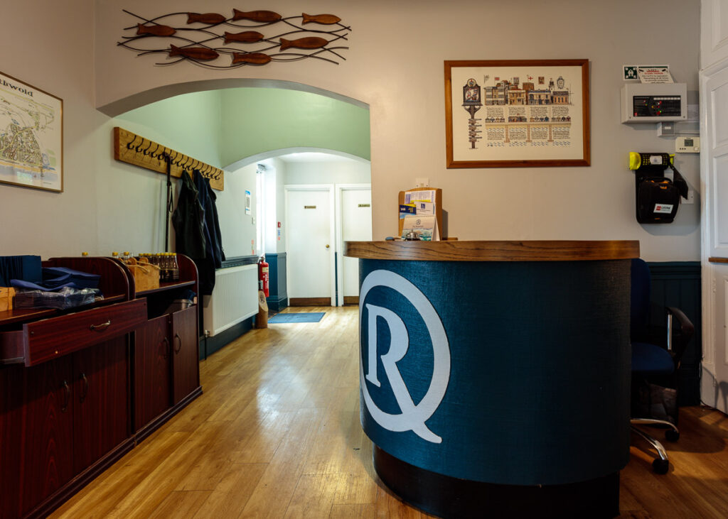 A hostel reception desk with a blue circular counter displaying a white R logo, wooden flooring, and nautical-themed decor throughout the space.