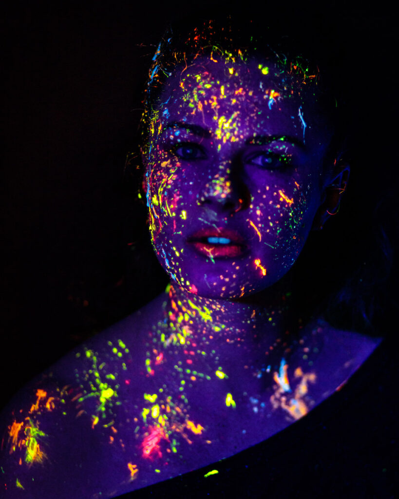 A person's head and shoulders covered in vibrant neon paint against a black background, glowing in shades of purple, yellow, blue, pink and orange.