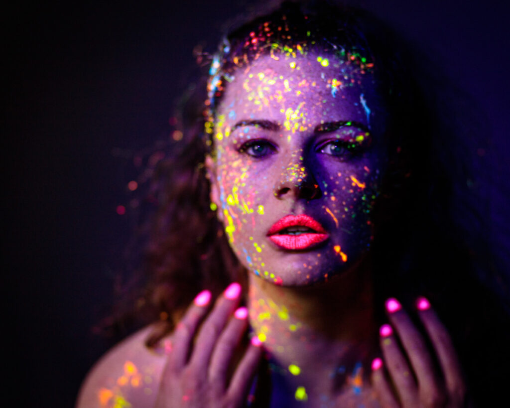 Woman's face covered in fluorescent paint particles against dark background, wearing bright pink lipstick and nail polish.