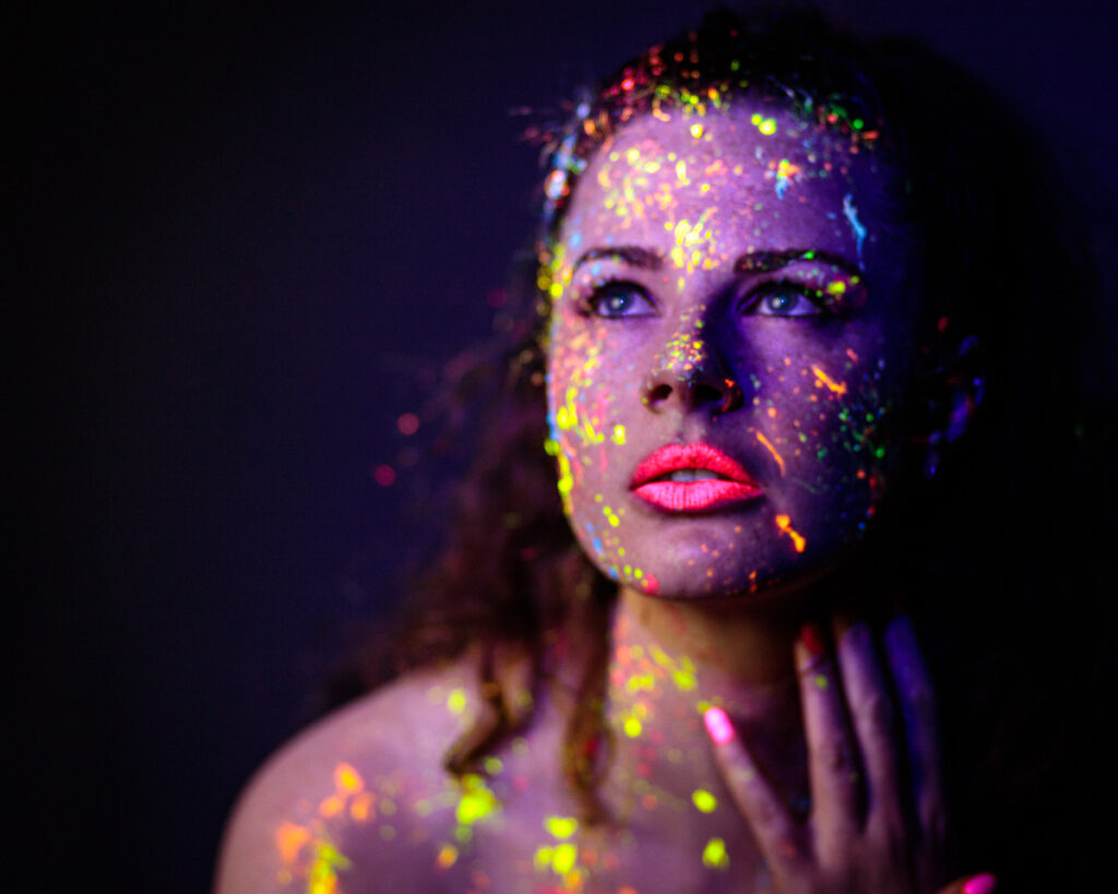 Woman's face covered in fluorescent paint against dark background, looking upward with striking eyes and bright pink lips.