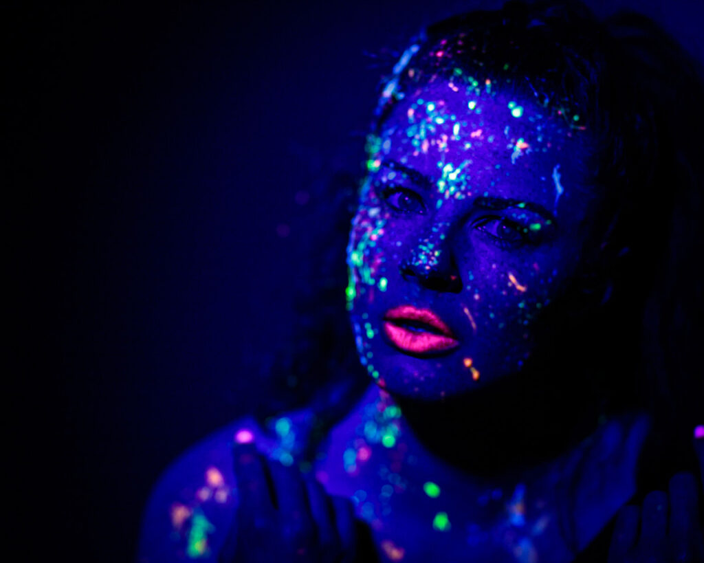 A person with neon body paint and makeup glowing under ultraviolet light against a dark background.