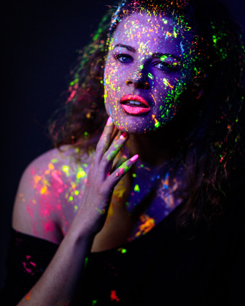 A woman's face and shoulders covered in vibrant neon paint against a dark background, touching her face with her hand.