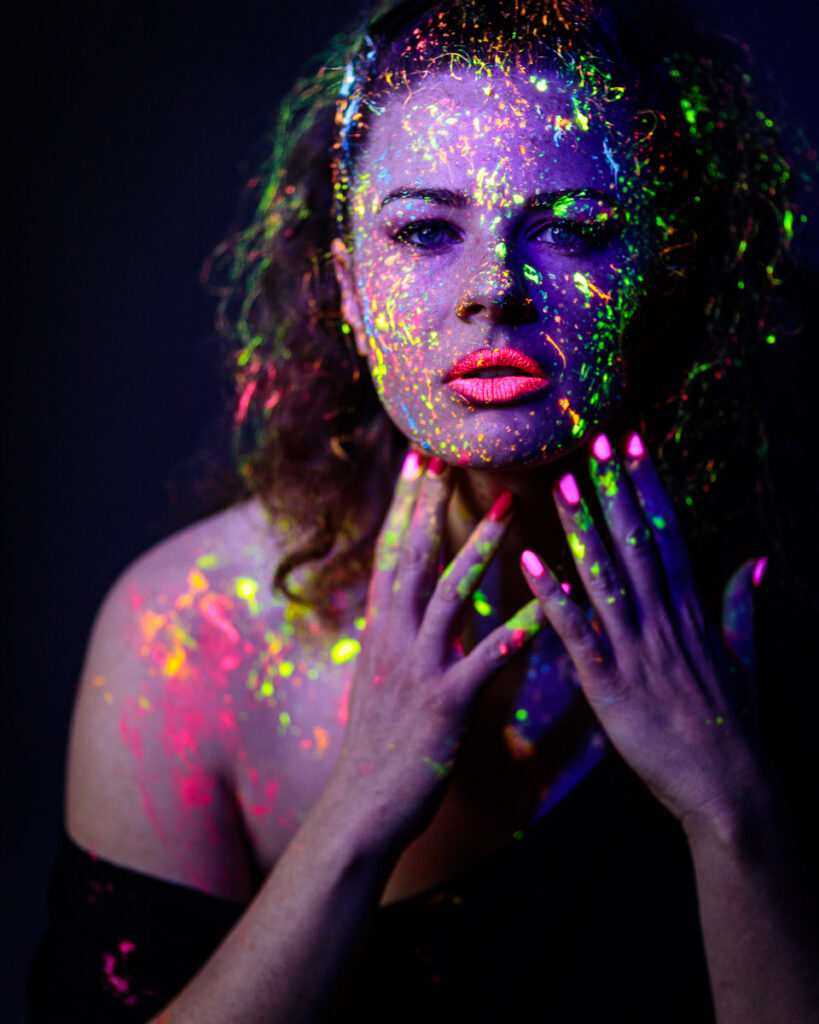 A woman's face and shoulders covered in vibrant neon paint against a dark background, looking directly at the camera.