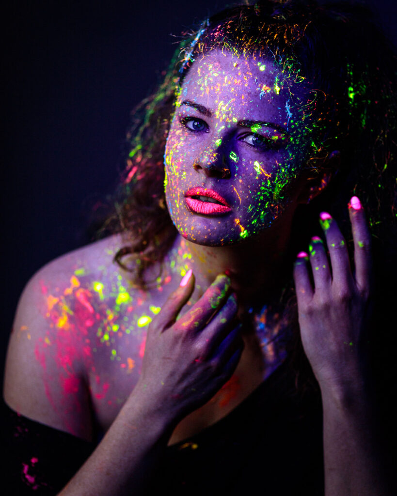 A woman's face and shoulders covered in vibrant neon paint in shades of green, yellow, pink and purple against a dark background.