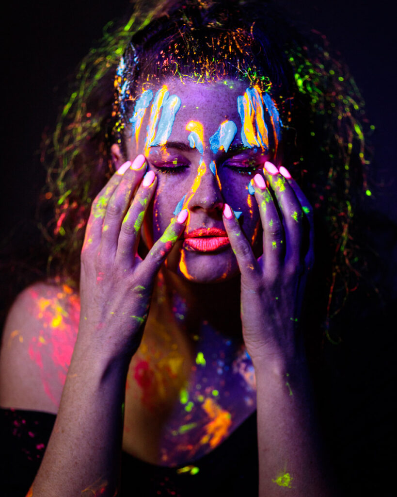 A person with fluorescent body paint and neon makeup holds their face with both hands against a dark background.
