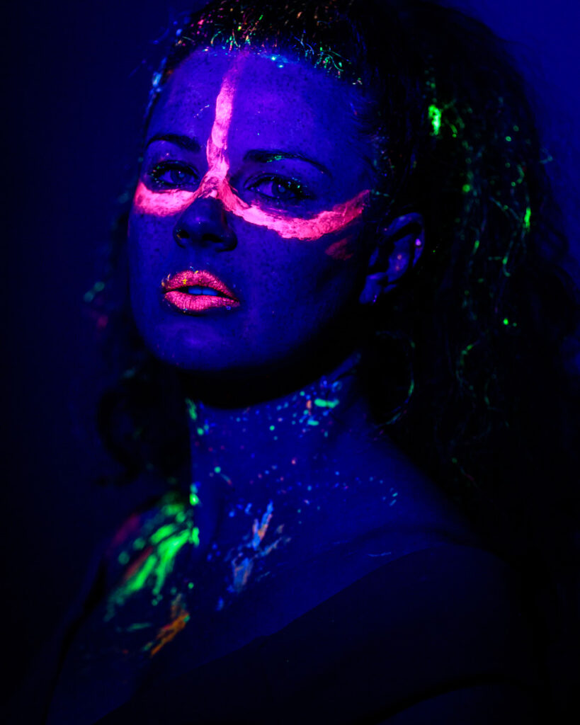 A person wearing neon face paint and glasses, illuminated under ultraviolet light against a dark blue background.