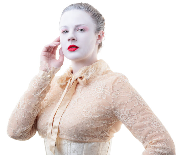 A woman with pale makeup and red lips wears a beige lace jacket over a cream top. She touches her face with one hand against a white background.
