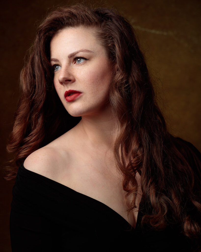 Portrait of Isabelle in an off-shoulder black top, long auburn hair, warm brown backdrop