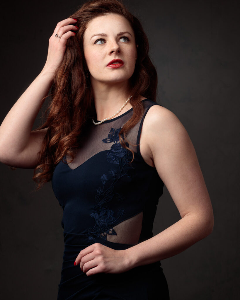Portrait of Isabelle in a navy dress with sheer panel, hand in her hair, dark background
