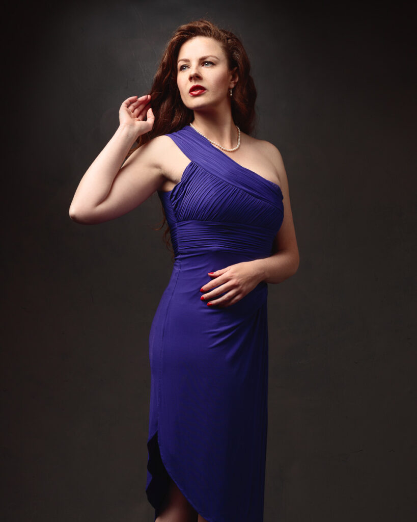 Portrait of Isabelle in a purple one-shoulder ruched dress, pearl necklace, dark background