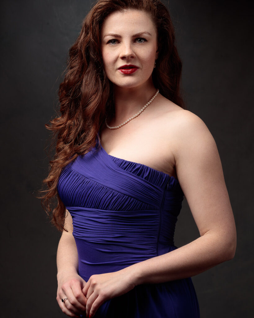 Close portrait of Isabelle in a purple one-shoulder dress, pearl necklace, dark background