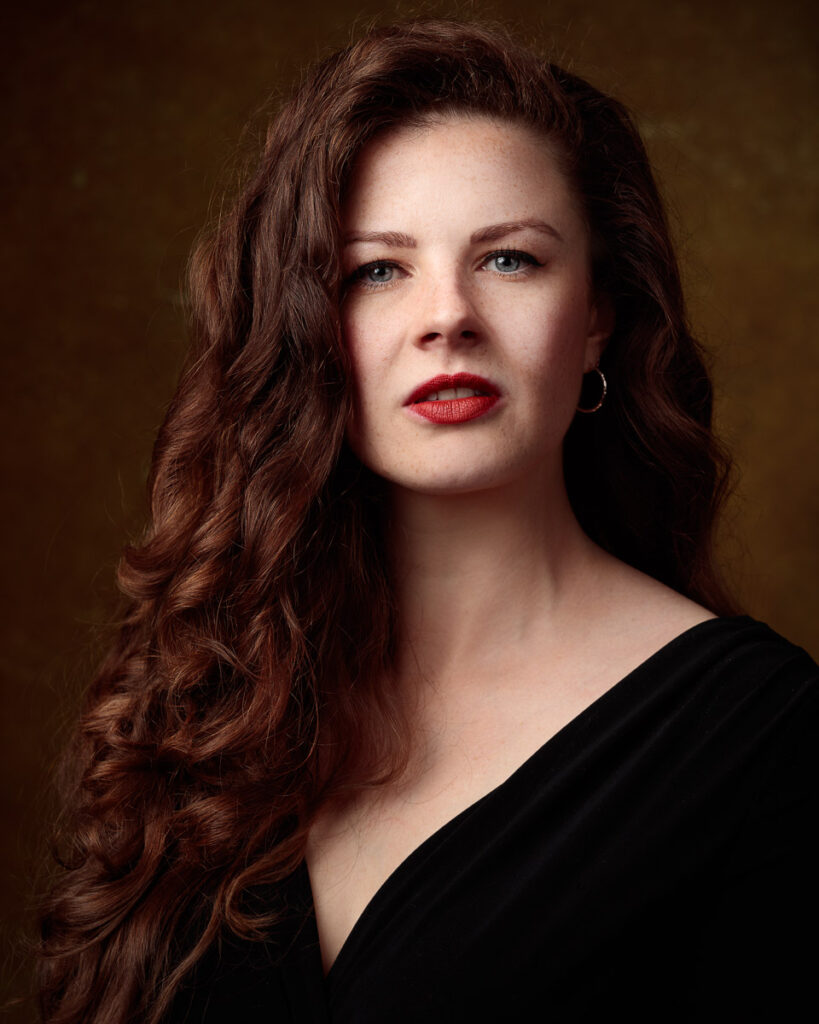 Portrait of Isabelle in an off-shoulder black top, warm brown backdrop, direct gaze