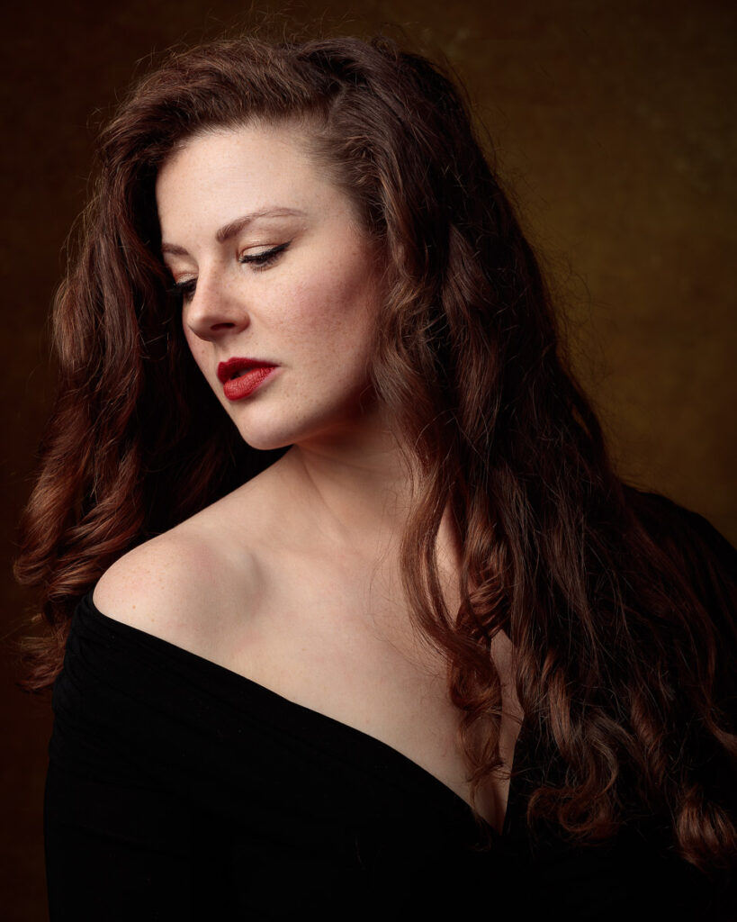 Portrait of Isabelle in an off-shoulder black top, looking down, warm brown backdrop