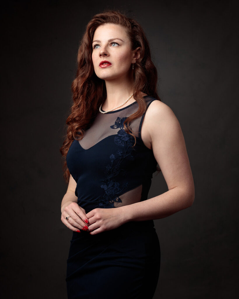 Portrait of Isabelle in a navy dress with sheer panel, pearl necklace, looking up, dark background