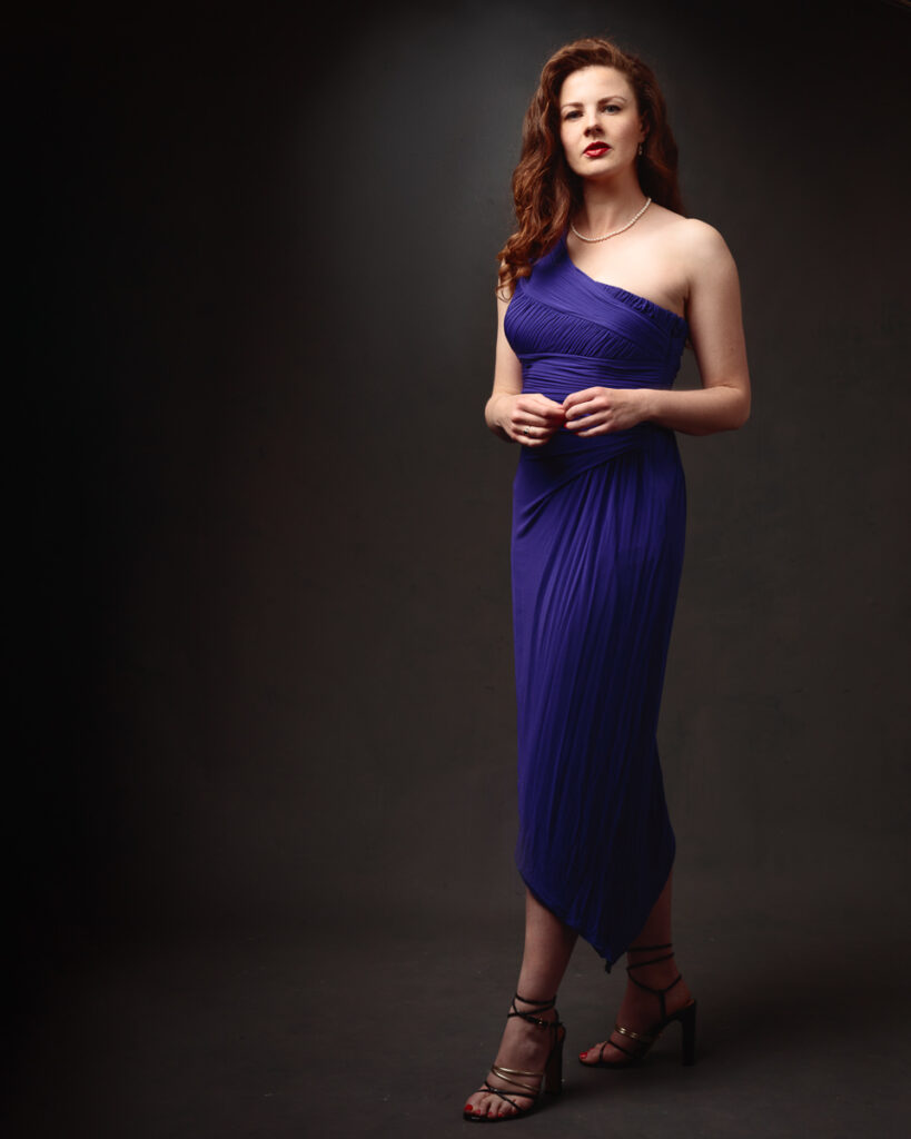 Full-length portrait of Isabelle in a purple one-shoulder dress, hands clasped, dark background
