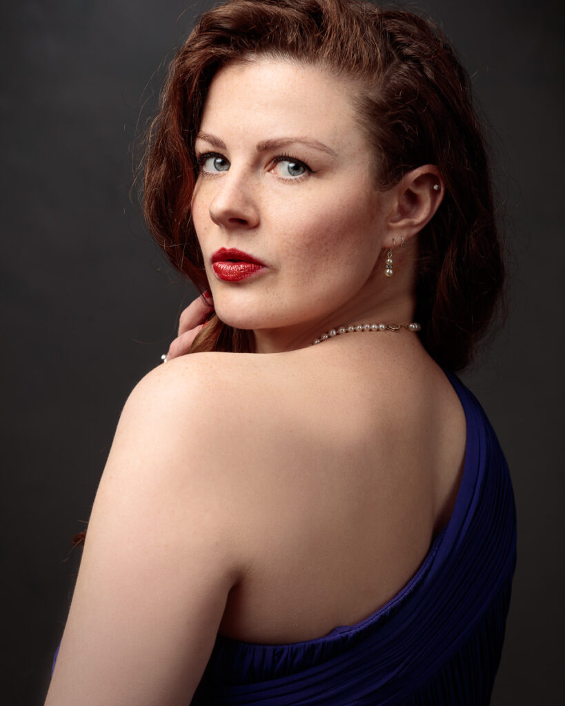 Close portrait of Isabelle looking back over her bare shoulder, purple dress and pearl necklace