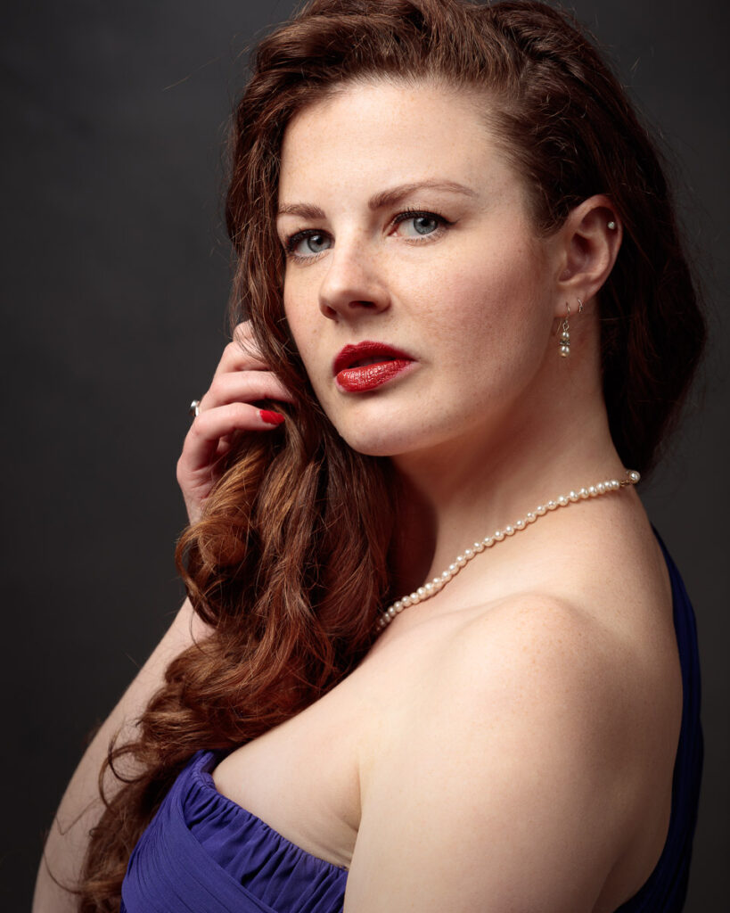Portrait of Isabelle in a purple dress and pearl necklace, hand touching her hair