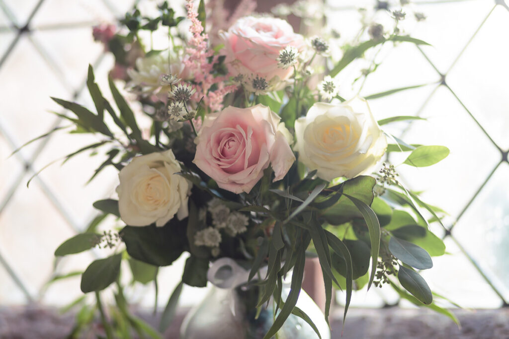 A floral arrangement with cream and pink roses, small white flowers, and green foliage in a vase, backlit by bright windows.